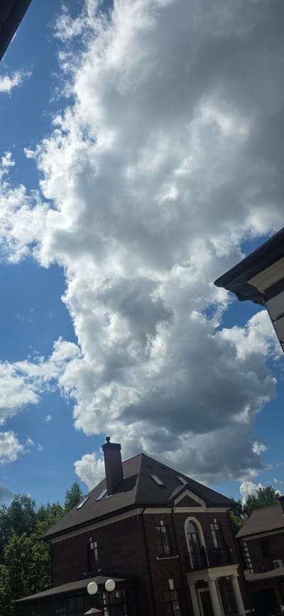 This media file features a low-angle view looking up at a partly cloudy sky, with elements of buildings visible. The sky is a vibrant blue, punctuated by large, fluffy white and grey clouds, suggesting a dynamic weather pattern. Two buildings are partially visible, framing the sky. On the left, a portion of a building with a white and brown facade is seen at an angle. On the right, a multi-story brick building with a dark roof occupies a significant portion of the frame, appearing to be modern architecture. This building has several windows, a chimney, and an air conditioning unit visible. Green trees are also seen near the base of the brick building, indicating an outdoor setting. The perspective is unusual, making it seem as if the viewer is lying down and looking straight up. The lighting suggests it is daytime, likely midday or afternoon, given the brightness of the sky and clouds. The location context provided is "Русская Деревня-2 Кп, Russia". There is no visible text in the image. The scene evokes a sense of looking up at the vastness of the sky and the surrounding architecture, with the clouds suggesting a pleasant but potentially changeable day.