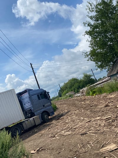 A blue semi-trailer truck with a white trailer is parked on a dirt road covered in wood chips and debris in Liubikha, Russia. The truck has "MAN G7H" written on its side. Overhead power lines run diagonally across the frame. In the background, there is a pile of logs, a rustic wooden building with a weathered roof, and dense green trees. The sky is blue with scattered white clouds. The scene appears to be an industrial or storage area.