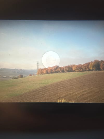The image displays a rural landscape identified as being in the city of Montepulciano, Italy, observed through a screen, indicated by dark borders at the top and bottom. The foreground features a large expanse of brown, tilled agricultural land with discernible furrows, transitioning to a strip of green grass on its left. A dense line of deciduous trees with vibrant autumn foliage (orange, yellow, brown) traverses the midground horizontally. To the far left, a tall, slender, grey metallic lattice tower, consistent with a telecommunications or power transmission structure, rises above distant hills and fields. Horizontal lines, possibly power lines, extend from the tower across the mid-distance. The background consists of rolling hills covered in a mixture of green and brown fields, receding into a light blue, hazy sky. A semi-transparent white circular overlay, featuring a central horizontal line and faint internal markings, obscures portions of the midground trees and sky in the upper-right quadrant, consistent with a graphical user interface element. No human figures or discernible actions are present.