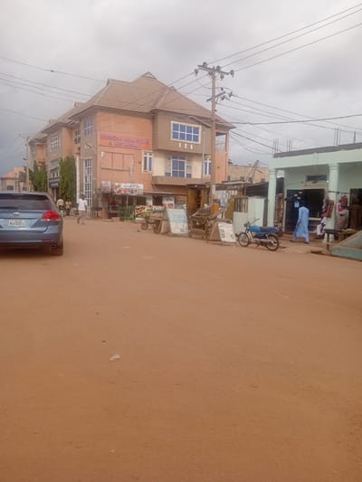 The image captures a street scene in Tundun Wada South, Nigeria, with a focus on a commercial building and its surroundings. A light blue sedan is parked on the left side of the dusty, reddish-brown road. The building in the center, painted in shades of orange and brown, features multiple stories with numerous windows. A prominent sign on the building reads "SEORORA MEGA STORE & RESTAURANT".
In front of the store, several people are visible. A man in a white t-shirt and gray pants is walking on the sidewalk, and another person is standing near a wheeled cart laden with goods. To the right of the main building, a light blue and black motorcycle is parked on the street. Further to the right, a light green building with pillars appears to be another shop. A person in a light blue robe is standing at the entrance of this green building, and another individual is seated nearby. Various items, possibly merchandise, are displayed outside the green building. Overhead, a utility pole with electrical wires crosses the sky, which is filled with cloudy conditions. The overall environment suggests a bustling commercial area on a typical day.