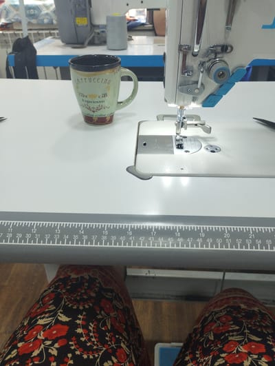 An overhead close-up shot shows a sewing machine with a measuring tape ruler in the foreground and a coffee mug on the left. The sewing machine is white with blue accents, and its needle is threaded with white thread. The mug is green with "CAPPUCCINO" and "Espresso" written in a vintage style, along with coffee-related imagery. The measuring tape ruler is grey and marked with black lines and numbers from 11 to 55. The background is slightly blurred, showing a table and what appears to be a serger or another sewing machine. The scene is set on a white tabletop. The image's perspective suggests it was taken from a seated position, looking down at the workspace. The presence of the sewing machine and the measuring tape indicates a sewing or tailoring environment. The context of Kyzyltog (Krasnogorsk), Uzbekistan, suggests this might be a workshop or a home-based sewing area within that city.