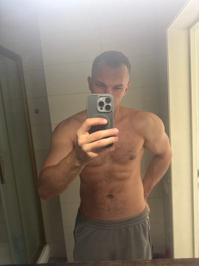 A shirtless man is taking a selfie in a bathroom mirror. He is wearing gray athletic shorts. The location is not discernible from the image.