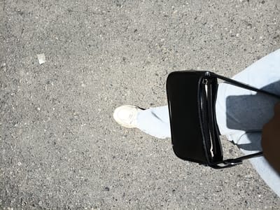 The image shows a person's leg, foot, and a black handbag on an asphalt surface. The person is wearing light blue jeans and white sneakers. The handbag is glossy black with a zipper closure and a strap. The asphalt is gray and speckled with small stones. A small white piece of litter is visible on the asphalt to the left of the foot. The lighting suggests it is daytime, with shadows indicating sunlight. The scene appears to be an outdoor urban environment, possibly a street or sidewalk in Osh, Kyrgyzstan.