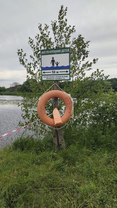 In the city of Ramon, Russia, a safety sign for a children's swimming area is prominently displayed on a young tree by the water. The sign, written in Russian, reads "Children's swimming area" and depicts an adult holding a child's hand by the water. Below this, it indicates a 50-meter zone and provides emergency contact numbers: "In case of accidents in the water, call 01, mobile 112." An orange lifebuoy hangs from the tree, with a small orange bag attached to it. A red and white striped caution tape is visible in the foreground, extending across the grassy bank toward the water. The scene is set against a backdrop of green foliage and a cloudy sky, with the calm water of a lake or river in the background.