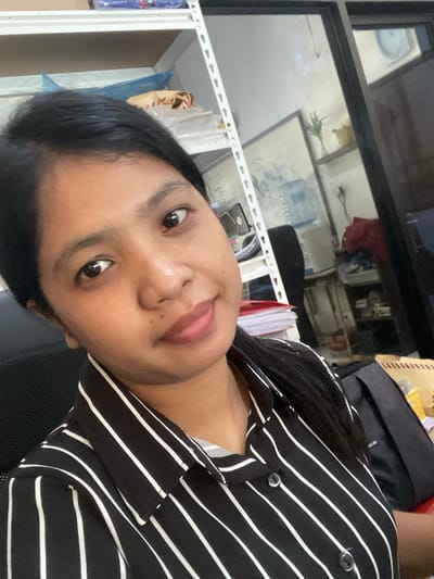 A medium-close-up indoor shot features a woman with dark hair wearing a black and white striped shirt. The background includes shelves holding various objects, office supplies, and a window. A dark bag is also partially visible. The location is potentially in Negara, Indonesia.