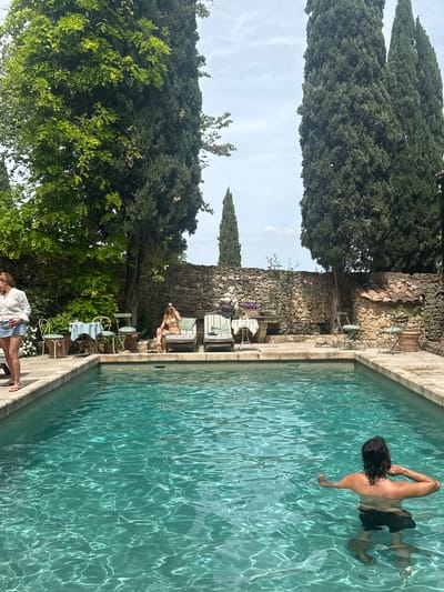 The image depicts an outdoor swimming pool with several people nearby. A man is swimming in the pool, facing away from the camera. A woman is sitting on a lounge chair next to the pool, and another woman is standing near the edge of the pool in the lower part of the image. The pool is surrounded by a stone wall, vegetation including tall, narrow trees and other plants and flowers. The setting appears to be in Saint-Victor-la-Coste, France, based on the style of the building and landscape. Chairs and tables are positioned along the edge of the pool, suggesting a relaxed or social setting.