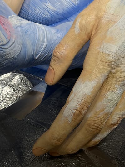 An oversized, hyper-realistic sculpted hand, rendered in a skin-tone hue and partially covered with white, paint-like residue, occupies the foreground. The hand features pronounced knuckles and detailed skin texture, with visible fingernails. It is positioned over a dark, subtly textured surface that includes a horizontal, reflective strip of material on the lower left. In the background, an undulating, ridged blue form, possibly drapery or another sculptural element, shows faint hints of red coloration. The scene depicts an art installation or sculpture exhibition within a venue in 성동구, South Korea. No human subjects are actively engaged in interaction with the sculpture.