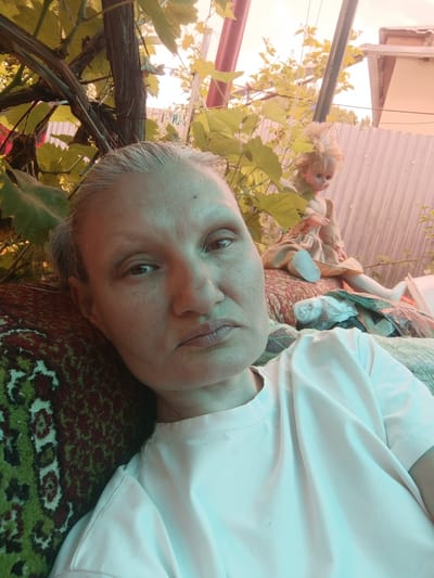A selfie taken outdoors in Ostrogorka, Russia, shows a middle-aged woman resting her head on a patterned cushion. She is wearing a white t-shirt and appears to be looking towards the camera with a neutral expression. Behind her, a porcelain doll with blonde hair is visible, sitting on what looks like another cushion or fabric. The background includes lush green foliage, a wooden pole, and a corrugated metal fence. The lighting suggests it is daytime, possibly late afternoon, with a warm, hazy glow. The overall setting appears to be a backyard or garden area.