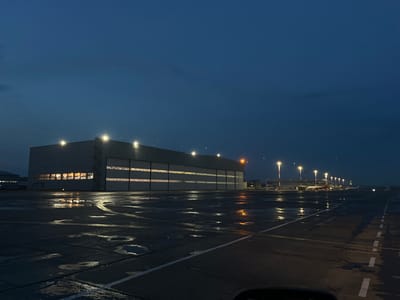 A large, modern hangar facility stands illuminated by lights both inside and out, with rows of large hangar doors visible. The facade of the building features a long horizontal band of windows showing warm light from within. Adjacent to the hangar, the tarmac is wet, reflecting the lights from the building and tall, pole-mounted lamps. In the distance, a few aircraft are parked near a terminal building, also illuminated. The sky is a deep twilight blue, indicating either early morning or late evening. The overall atmosphere is quiet and serene, with the reflections on the wet surface creating a sense of depth and stillness. The location appears to be an airport, likely Yemelyanovo Airport in Russia, given the context provided. There is no visible text in the image.