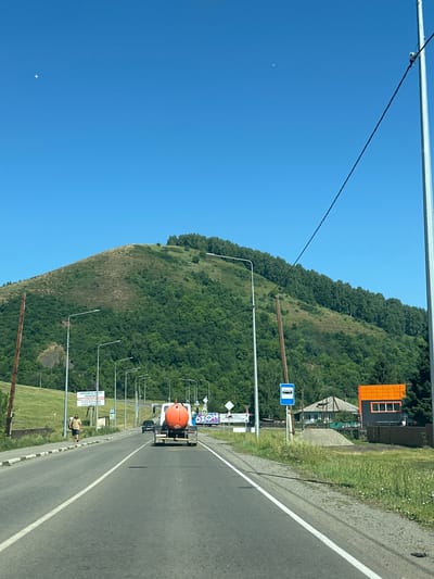The image shows a roadside scene in the city of Aya, Russia, on a clear, sunny day. A two-lane asphalt road runs horizontally across the frame, with white lane markings visible. In the foreground, on the right side of the road, a tall, metallic pole supports a black wire that stretches diagonally across the sky. A blue bus stop sign is mounted on a pole on the right side of the road.
Several streetlights line the left side of the road. A pedestrian walkway with a curb is visible along the left edge of the road. A man wearing a striped shirt and shorts is walking away from the camera on the walkway.
Further down the road, a truck with an orange cylindrical tank is traveling away from the camera. In the background, a large, tree-covered hill dominates the landscape. Some houses and buildings are visible on the right side of the road, with an orange-tiled roof on one of them. A billboard with Cyrillic text is also present near the left side of the road. The sky is a clear, bright blue with a small crescent moon visible in the upper left quadrant of the image. The environment appears to be rural or semi-rural, with a mix of natural elements and infrastructure.
