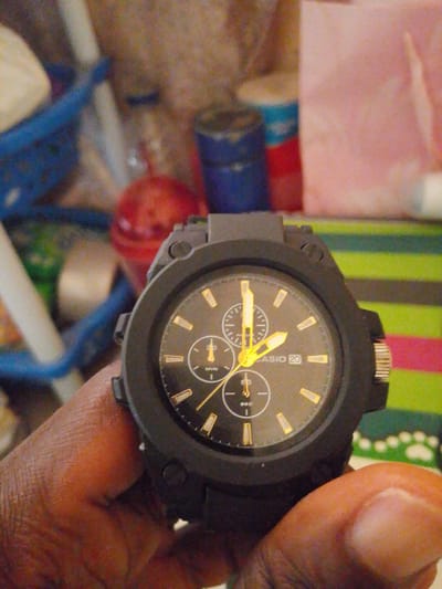 A close-up, slightly blurry, eye-level shot shows a black Casio watch with gold accents on a person's wrist. The watch face displays the brand "CASIO 20", hour markers, and three sub-dials. The person's hand, with visible skin texture and subtle red staining on the palm, is holding the watch. In the background, out of focus, are various colorful items including what appears to be a blue plastic basket, red and blue cylindrical objects, and a pink textile. The lighting is moderate. The location is identified as Abeere, Nigeria.
