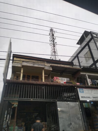 A telecommunications tower stands tall against an overcast sky, situated behind a multi-story building with a yellow facade. The building has multiple balconies, one of which has laundry hanging out to dry. A modern building with a black and white geometric design is visible to the right of the telecommunications tower. Below the yellow building, a storefront with a metal gate is visible, displaying a sign that reads "MENJUAL: SNACK." A person wearing a black t-shirt and a cap is walking away from the camera in front of the storefront. The environment appears to be a densely populated urban area in Gunungbukit, Indonesia, indicated by the presence of multiple buildings, power lines, and commercial activity.