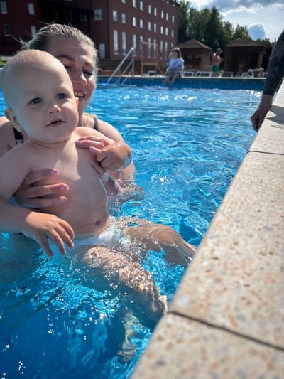 A woman is holding a baby in a swimming pool. The baby is facing the camera and has blue eyes. The woman is smiling and her hair is wet. There are other people in the background, some sitting on lounge chairs and others in the pool. There is a building in the background with many windows. The pool area appears to be part of a resort or hotel. The location is specified as the city of Yubileynyy, Russia.
