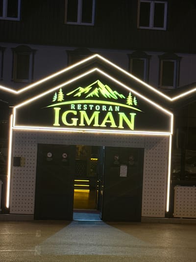 A neon sign displaying "RESTORAN IGMAN" is illuminated above the entrance to a building. The sign features a green mountain graphic with pine trees. The entrance itself has two large black doors. The building is lit with white neon lights forming a decorative outline around the sign and entrance. The ground in front of the building appears wet, reflecting the lights. The overall scene depicts the exterior of a restaurant at night.
