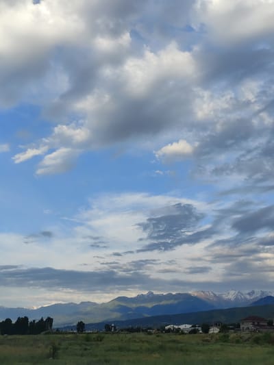 The image shows a landscape scene in Dolan, Kazakhstan. A wide expanse of sky, filled with a mix of white and gray clouds, dominates the upper half of the frame. The clouds are varied in shape and density, with some appearing wispy and others more solid. The lower half of the image features a view of distant mountains, their peaks capped with snow. The mountains are a deep blue-green, with patches of sunlight illuminating some of the higher slopes. In the foreground, a grassy field stretches across the bottom of the image, with scattered trees and a few buildings visible on the horizon. The buildings appear to be houses, with one having a dark red roof. A water tower is also visible. The overall impression is of a natural landscape under a dramatic sky, suggesting either early morning or late afternoon due to the lighting on the mountains. The weather appears to be partly cloudy, with breaks of blue sky visible between the clouds. No people or specific activities are depicted. There is no visible text in the image.
