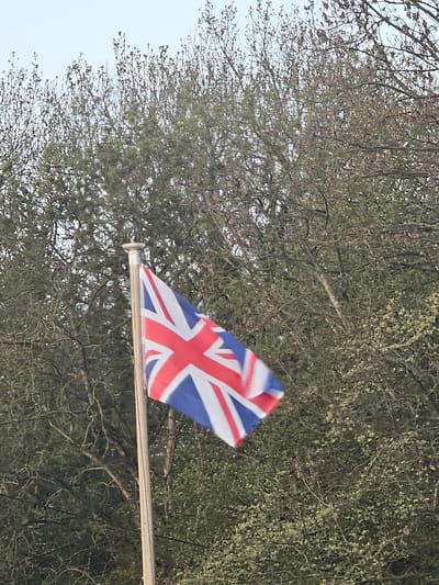 The image shows a Union Jack flag waving on a pole against a backdrop of trees with budding leaves in Holywell, United Kingdom. The sky is partially visible in the upper left corner.