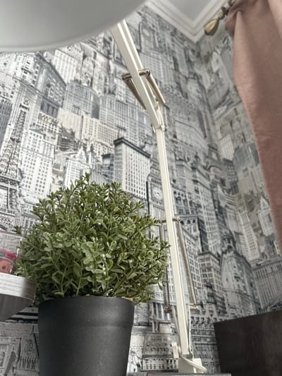The image depicts a close-up view of a desk or table surface with several objects. A small, artificial green plant in a black pot sits on the surface, slightly tilted. A white, articulated lamp with visible springs extends across the frame. The background is wallpapered with a black and white cityscape pattern. A section of a peach-colored curtain is visible in the upper left corner. A dark-colored shelf or surface is present in the upper right corner.