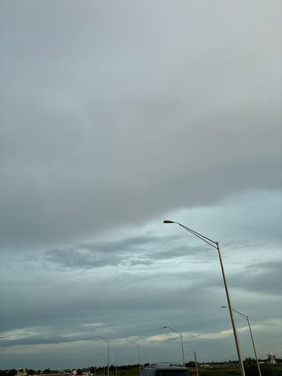 An overcast sky fills the upper portion of the image, with layers of grey and muted blue clouds stretching across the frame. Several tall streetlights with metallic poles and single light fixtures are spaced along a road in the lower portion of the image. The road itself is lined with trees and some buildings in the distance. There are vehicles visible on the road, including a dark SUV in the foreground and several other cars further down. The scene appears to be a highway or a major road in a suburban or developing area. The overall impression is one of an approaching storm or a cloudy day. This image was captured in Lakewood Ranch, United States.