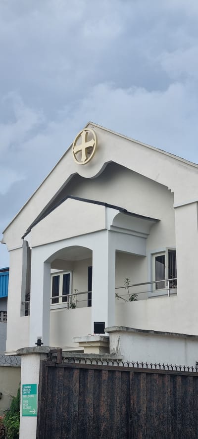 The image shows the exterior of a building in Ijaiye, Nigeria. The building is modern with white and grey architectural elements, including balconies with metal railings and several windows. A distinctive gold circular emblem with a cross design is affixed to the building's facade near the top left. To the right of the building is a large, dark metal gate with decorative spikes along the top. A green sign is attached to the wall near the gate, with white text that reads: "LONGONG MEDICAL CENTRE WELCOME THIS WAY PLEASE". The sky is overcast, indicating a cloudy day. The perspective of the photo is from a low angle, looking upwards towards the building and sky, with the image rotated 90 degrees clockwise.