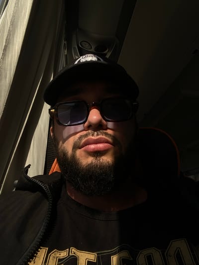 This is a close-up, low-angle selfie of a person wearing sunglasses, a black beanie, and a black jacket over a black t-shirt with large, yellow, distressed lettering. The person has a beard and is looking towards the camera. The lighting suggests it is daytime, with a strong light source creating dramatic shadows across the person's face and the surroundings.

The setting appears to be indoors, possibly in a vehicle or a room with a window. Visible in the background is a curved ceiling panel, a ventilation grate, and some fabric that could be a curtain or upholstery. The overall mood is somewhat enigmatic due to the strong shadows and the person's obscured eyes. There is no clear indication of the specific activity or event taking place, beyond the act of taking a selfie.

The visible text on the t-shirt is partially obscured by shadow but appears to be a large, stylized word or logo. On the beanie, there is a visible emblem with text that is difficult to decipher due to its small size and the image quality. There are no other discernible text elements in the image. The image is oriented sideways, with the person's head at the top.