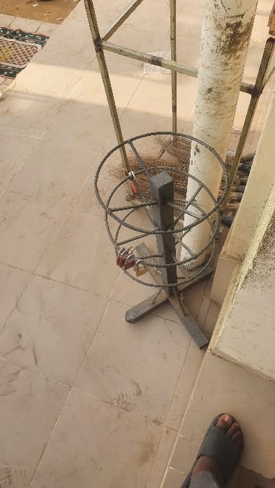 This image depicts an outdoor tiled area in Danbare, Nigeria, illuminated by daylight. The ground is composed of dirty, light-colored square tiles with visible grout lines.

A dark-colored metal stand, comprising a central vertical rectangular post and two circular, spoked metal frames, is centrally located. Multiple padlocks, including two reddish-brown and one metallic, are affixed to the upper circular frame. Behind the stand, a dark-bristled broom is partially visible.

To the right, a stained, off-white cylindrical pole stands adjacent to a light-colored wall or pillar. In the upper left and central background, a lighter, possibly yellowish-green, metal support structure constructed from welded square tubing is partially visible. The upper left corner also shows a fragment of a green, yellow, and white patterned mat on a dirt surface beyond the tiles. In the lower right foreground, a human foot, wearing a dark grey slide sandal, is partially visible.