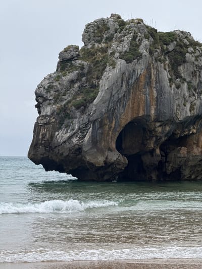 The image captures a striking geological formation, a large, rugged rock outcrop, jutting out from the sea. The rock is textured and layered, with visible striations and patches of green vegetation clinging to its surfaces. A notable feature is a dark, cavernous opening or arch carved into the rock, creating a dramatic contrast with the lighter stone. The sea laps gently against the base of the rock, with small waves breaking and creating a foamy white edge where they meet the sandy shore. The water is a muted green, reflecting the overcast sky above. The beach appears to be a mix of sand and small pebbles. The overall scene is one of natural beauty and rugged coastal landscape. The time of day appears to be overcast, as indicated by the soft, diffused light and the gray tones of the sky. The location is identified as Llanes, Spain, suggesting a coastline characteristic of this region of northern Spain. There is no text visible in the image.