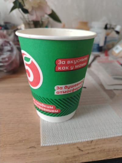A green and white paper cup with red lettering is placed on a wooden table with a textured placemat underneath it. The cup has text in Russian, including "За вкусным, как у мамы" (For delicious, like mom's) and "За душевной атмосферой" (For a soulful atmosphere). There is also text that says "сладким настроением" (with a sweet mood). The background shows blurred items, including what appears to be flowers in a vase and some packaging. The overall scene suggests a casual setting, potentially a cafe or home environment in Aramil, Russia, given the Russian text on the cup. No people are visible in the image.