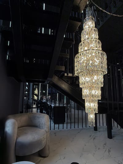 A low-angle interior shot captures a dimly lit space with a prominent, multi-tiered crystal chandelier hanging from the ceiling. The chandelier features several cascading layers of faceted crystals, reflecting and refracting the warm light emanating from within. To the left of the chandelier, a plush, light-colored armchair with rounded arms and a circular seat is visible. The armchair is positioned on a floor with a marble-like pattern. Behind the armchair and to the right, a dark, industrial-style staircase with metal railings ascends. The staircase appears to be constructed from black metal beams and treads. In the background, through the open structure of the staircase and beyond a railing, glimpses of other areas of the establishment are visible, including tables, chairs, and more vertical lighting fixtures. The overall ambiance suggests a modern, somewhat luxurious, and possibly dimly lit establishment. There are no explicit details that would indicate the location is in Укромновское сельское поселение, Ukraine.