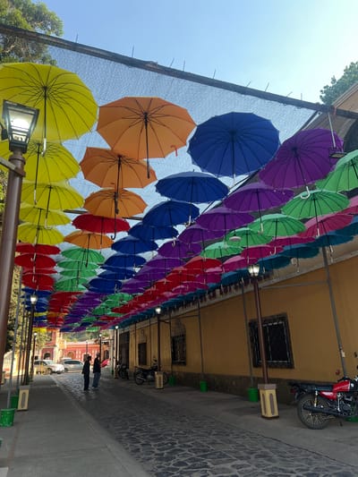 An eye-level outdoor shot captures a street in La Esperanza, Honduras, covered by a canopy of colorful umbrellas. The umbrellas, ranging in shades from yellow to purple, are suspended above a walkway that runs along the side of a building. Two figures are standing in the background. The walkway is constructed from cobblestones. A motorcycle is parked next to the building to the right.