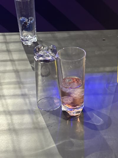The image shows three horizontally aligned glass tubes. The top tube contains a brown liquid and ice cubes. The middle tube appears to be empty and clear. The bottom tube contains a dark blue substance. The tubes rest on a gray surface. The background includes a blue light and a dark blue diagonal pattern. The location in the city of Dartford, United Kingdom is unknown.