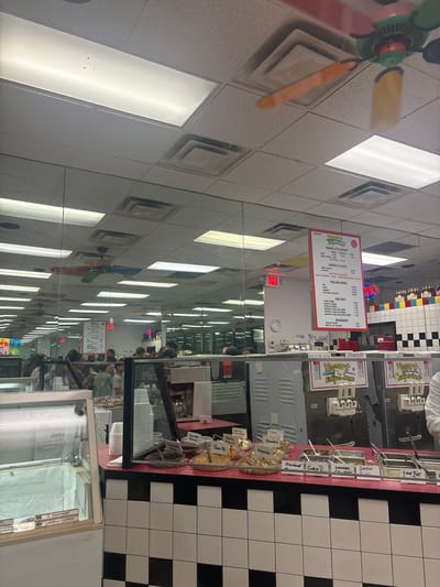 The image shows the interior of Yogurt Emporium, a yogurt and ice cream shop. The storefront is located in Boca Raton, Florida. A colorful menu board is visible with pricing options for yogurt, ice cream, shakes, floats, pies, and beverages. A series of frozen yogurt machines are lined up along a countertop with various toppings such as graham crackers, granola, almonds, and chocolate chips, in transparent containers. Customers are visible in the reflection of the glass, suggesting an active business environment.
The interior design includes a black and white checkered wall pattern. A ceiling fan is also visible.