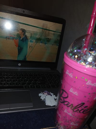 The image shows a laptop partially obscured by a pink Barbie-themed cup with a straw and glitter inside. The laptop screen displays a person playing a sport within a net-enclosed area. The keyboard of the laptop is visible, with a sticker of a woman on the right side. An HP logo is visible on the laptop's body. A dark blue surface is partially seen in the bottom right corner of the image. There is no information to determine the location in the city of Salar, Uzbekistan.