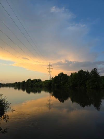 The image depicts a serene sunset over a wide river in the city of Togashevo, Russia. The sky is a gradient of colors, transitioning from a pale yellow and orange on the horizon to a soft blue overhead, with scattered clouds. Several power lines stretch diagonally across the sky, leading the eye towards a tall electricity pylon situated on the far bank. The river's surface is calm, perfectly reflecting the sky, the pylon, and the dense line of green trees that borders the opposite bank. On the left bank, some vegetation and branches are visible close to the water's edge. The overall atmosphere is peaceful and tranquil, characteristic of a natural landscape at dusk. There are no people or animals visible in the scene.