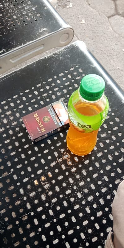 The scene, located in Bojongherang, Indonesia, presents a static, elevated view of objects resting on a black, perforated metal table. A red and black pack of "MAGNUM MAX" cigarettes, labeled "20 KRETEK FILTER," is present. Adjacent to the cigarette pack is a clear plastic bottle containing an amber-colored liquid, identified as "Frestea" with a green cap bearing "Product of The Coca-Cola Company" text. Condensation is visible on the bottle's exterior. A partial view of a human leg or knee, clothed in light-colored fabric, is visible in the lower right foreground. The background reveals light-colored, patterned paving or concrete ground and a section of another similar perforated metal structure.