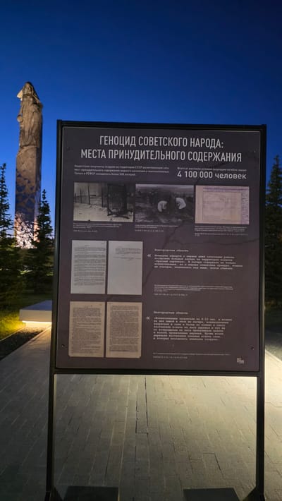Here is a detailed description of the image:
**Scene Description:**
The image shows an information panel standing in an outdoor location, likely a memorial site or museum. The panel is upright and displays text and images related to the "Genocide of the Soviet People: Places of Forced Detention." The panel's text is in Russian, detailing the Nazi occupation's establishment of a network of forced detention sites across the USSR. The panel mentions over 500 camps in the RSFSR alone, with over 4,100,000 people perishing due to harsh occupation conditions. There are images of camps and historical documents.
The location appears to be outdoors, with brick pavers visible on the ground. Behind the panel, there's a statue in the background, partially obscured by trees. The sky is a gradient of blue, suggesting dusk or dawn. There is artificial lighting illuminating the pavers on the right side of the image.
**Location:**
Based on the text and subject matter, this is likely a memorial or museum in or related to Zaitsevo (Verevskoe S/P), Russia. The presence of a memorial statue further confirms that it is a dedicated memorial location.
**Objects:**
* Information Panel
* Brick pavers
* Statue
* Trees
* Artificial lights
* Historical Documents
**Overall Impression:**
The scene creates a somber atmosphere, highlighting a dark period in history. The erected panel and statue suggest commemoration and remembrance.