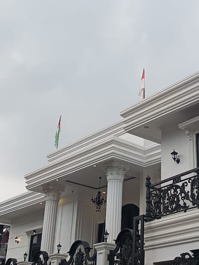The image presents a low-angle, exterior view of a large, neoclassical-style building in Sindangasih, Indonesia, beneath an overcast sky. The structure is predominantly white, featuring multiple fluted columns with ornate Corinthian capitals supporting an expansive, multi-tiered cornice.

Two flags are prominently displayed from the roofline: a flag with vertical stripes of red, white, and green is visible on the left, and a flag with horizontal stripes of red over white is on the right. An ornate black wrought-iron railing, indicative of a balcony or terrace, is visible on the upper right portion of the building. Exterior lighting includes several black, antique-style wall-mounted lanterns and recessed downlights embedded in the ceiling overhangs. A black chandelier with multiple arms hangs within a portico, and a small security camera is mounted discreetly near one of the columns. Dark-framed windows or doors are partially visible. No individuals or discernible activities are present in the scene.