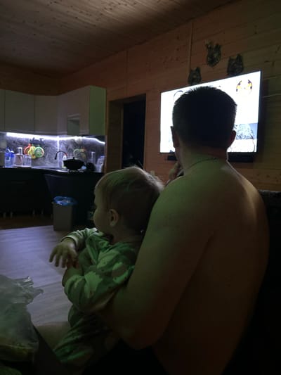 The image shows an indoor scene, likely a home interior. A shirtless man is seen holding a young child. The child is wearing a patterned shirt. In the background, a television is mounted on a wooden wall. Below the television, there appears to be a display case with illuminated shelves holding various objects. The location is identified as Bol'shakino, Russia.