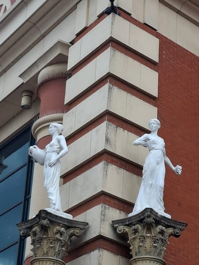 The image shows a two statues of women standing on ornate columns against a building with red brick and cream colored stone accents. The statue on the left is a woman holding a large jug. The statue on the right is a woman holding a bunch of grapes. The building has large windows and architectural details like cornices and pilasters. The sky is overcast. This is a photograph of the statues on the exterior of the Imperial War Museum North, located in Trafford, United Kingdom.