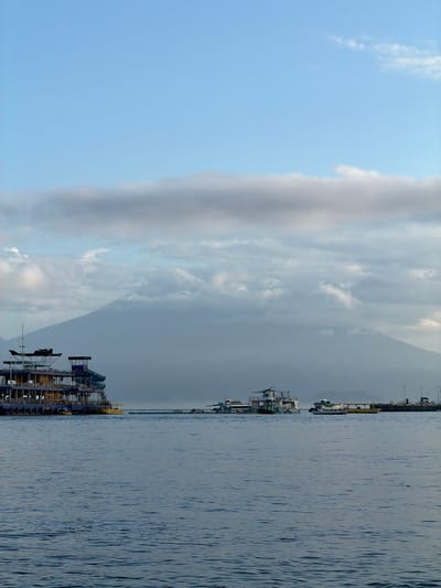 The image shows a portion of the cityscape of Toyapakeh, Indonesia. The scene encompasses a body of water, likely a harbor or bay, with boats of various sizes docked along the coastline. The sky is dominated by clouds, with some areas of blue sky visible. In the background, a mountain or large hill is partially obscured by the clouds.
The overall scene is one of a typical coastal town with maritime activity. The presence of boats suggests fishing or tourism activities. The weather appears to be overcast. The mountain provides a natural backdrop to the urban landscape.