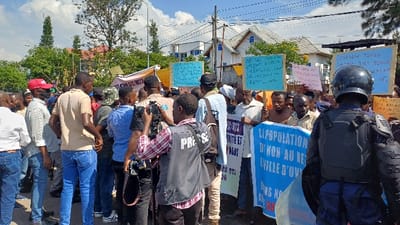 The image depicts an outdoor daytime gathering of approximately 30-40 individuals, primarily adult males, on a street in Kabinzi, Rwanda. The assembly appears to be a public demonstration or protest. Multiple participants are holding aloft light blue rectangular signs featuring handwritten text in an indigenous language. A prominent blue banner in the foreground displays partial text in French, including "LIPOPULATION DI NON AU RE" and "VILLE D'UV." A media representative, identifiable by a grey vest with "PRESS" emblazoned on the back, is visible in the mid-ground operating a video camera. Other individuals are also present with recording devices. In the immediate right foreground, a police officer, observed from the rear, is equipped with dark riot gear, comprising a helmet with a visor, chest and shoulder protection, and a clear riot shield. The environmental backdrop includes multi-story buildings with light-colored exteriors and dark roofs, interspersed with mature trees and overhead utility lines against a sky with scattered clouds.
