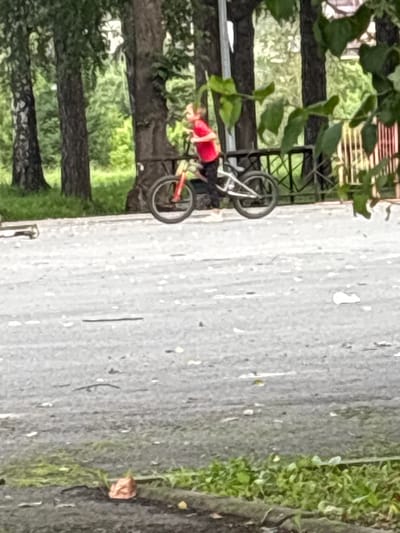 In Nevyansk, Russia, a person wearing a red shirt and black pants is riding a bicycle on an asphalt path. The bicycle has red handlebars and silver frame components. Behind the cyclist, trees with dark bark and green foliage are visible. The ground is covered in a mixture of asphalt and patchy green grass. Leaves are scattered on the ground.