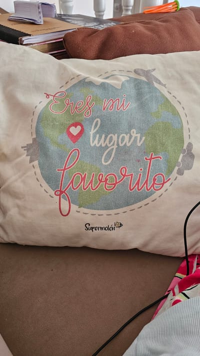 This is a close-up, eye-level shot of a cream-colored tote bag with a graphic design of a world map. The world map is stylized with green and blue continents, and a dotted line circling it, resembling a travel route. There are small, grey icons of a car and an airplane on this route, suggesting travel.

Dominating the graphic are two lines of text in a flowing, cursive font. The first line, in pink, reads "Eres mi" (You are my). Below this, in a larger, bolder red font, is "lugar favorito" (favorite place). A small pink heart with a dotted outline is placed between "mi" and "lugar." To the right of the graphic, near the edge of the bag, is a small, black logo with a stylized bee and the word "Supermolón" written below it in cursive.

The bag is resting against a brown, textured surface, possibly a sofa or a piece of furniture. To the left of the bag, part of a brown pillow with an orange object clipped to it is visible. The background is a bit cluttered with a few books or notebooks and some white railings. The lighting suggests it is daytime, likely indoors. The overall mood conveyed by the graphic is one of affection and a love for travel. The location context provided is María De Huerva, Spain.