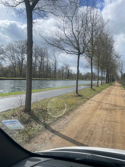 The image depicts an outdoor, static scene in Geel, Belgium, captured from the interior of a vehicle looking forward. The lower portion of a white vehicle's dashboard is visible, and a light-colored sticker with blue and green elements is affixed to the lower-left of the windshield.

On the left, a wide canal with dark, rippled water extends into the background. A narrow, wet, paved path runs parallel to the canal's near bank. To the right of this path, a row of uniformly spaced, bare-branched deciduous trees stands. These trees cast shadows to the right onto a wide, unpaved dirt road, which also extends into the background. The dirt road exhibits visible tire tracks and scattered gravel. Beyond the canal, on its far bank, a dense line of bare deciduous trees is visible. The sky above is partly cloudy, featuring prominent white and gray clouds. No people or animals are present.