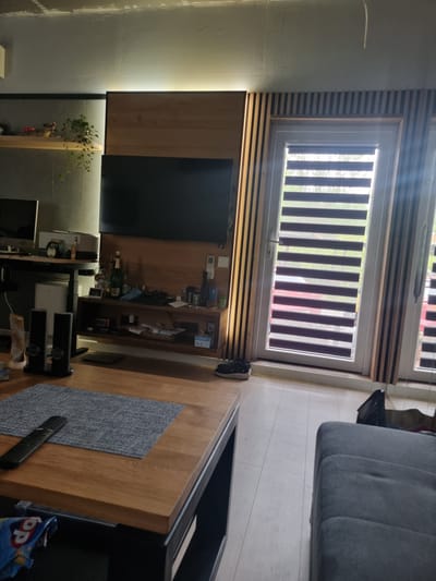 The image depicts an interior view of a living room, likely in a modern apartment or house. The primary focus is on a wall-mounted entertainment unit featuring a large black television and several wooden shelves below it. These shelves hold various items, including bottles, a framed picture, and decorative objects. To the left of the entertainment unit, there is a desk with a computer monitor and other office supplies. In the foreground, a wooden coffee table with a grey placemat and a remote control is visible. A portion of a grey sofa is seen on the right side of the image. A large window with horizontal blinds covers most of the right wall, allowing natural light to enter the room. The flooring appears to be light-colored wood. The overall ambiance is contemporary and tidy. There are no people or animals present in the scene. The location is identified as Szczaki, Poland.
