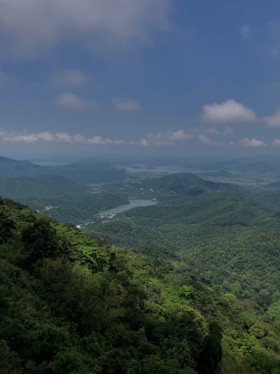 This is a high-angle, wide shot of a lush, mountainous landscape with a body of water in the distance, likely taken from a viewpoint in Nam Hải, Vietnam. The foreground is dominated by a steep, verdant mountainside covered in dense green foliage, with some trees casting shadows. The midground reveals rolling hills covered in a similar thick forest. In the distance, a winding river or inlet, with some structures visible along its banks, flows through the valley. Beyond the immediate hills, a series of progressively more distant mountain ranges fade into a hazy, atmospheric perspective under a blue sky dotted with scattered white clouds. The scene suggests a natural, undeveloped area with evidence of some human habitation and infrastructure, such as roads and buildings, integrated into the landscape.