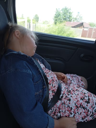 The image shows a young girl sleeping in the back seat of a car. She is wearing a denim jacket over a floral dress with pink and white flowers. A black seatbelt is visible across her chest and lap. Her head is resting on the car's headrest, and her eyes are closed, indicating she is asleep. Her blonde hair is pulled back in a ponytail.
The scene outside the car window suggests it is daytime, with a bright sky and green trees. There are some buildings and a wooden fence visible in the background, hinting at a rural or suburban location. The overall impression is one of a comfortable journey. No visible text is present in the image. The context provided places the scene in Bol'shoi Bereznik, Russia.