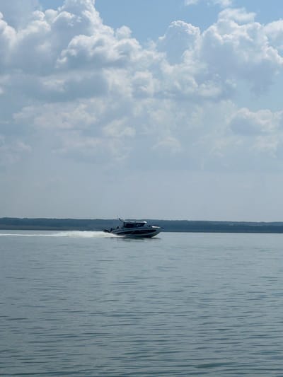 A motorboat is depicted traveling across a body of water, creating a white wake behind it. The boat is dark in color with white accents and appears to have several occupants. In the background, a dark, tree-lined shore is visible, suggesting a natural or semi-rural setting. The sky above is a pale blue, partially filled with fluffy, white cumulus clouds. The water is calm with subtle ripples. The image does not provide any specific visual cues to pinpoint its location to Bystrovka, Russia.