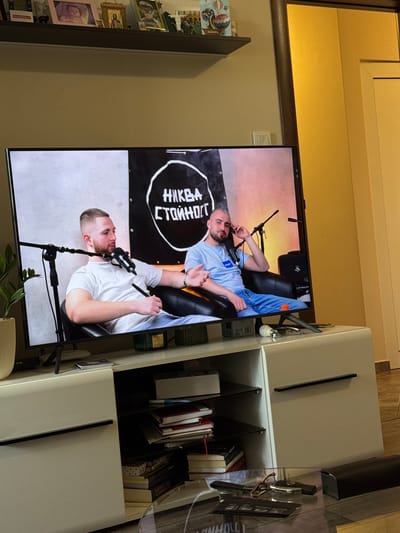 The image shows a television screen displaying a podcast or talk show with two men sitting in chairs, each with a microphone in front of them. A black banner with white Cyrillic text hangs behind them. The television is placed on a white media console. On the shelf above the television, several framed pictures and decorative items are visible. The media console itself contains shelves with books and other assorted items. In the foreground, a glass coffee table holds keys and other small objects. The overall scene appears to be an indoor living space or entertainment area. The specific location of Gara Elin Pelin, Bulgaria, is not discernible from the visual information provided in the image.