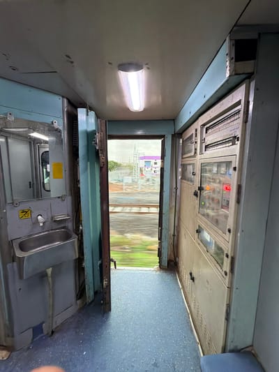 The image shows the interior of a train carriage, likely in Saphale, India. In the foreground, a blue-carpeted floor is visible. To the left, there is a grey sink with a faucet and a soap dish mounted on a blue panel. Above the sink, a mirror reflects part of the carriage. Adjacent to the sink, a yellow caution sticker is visible. To the right, a large, beige electrical panel runs along the wall, featuring multiple compartments with buttons, switches, and indicator lights, some of which are illuminated. The open doorway in the center of the image offers a view of the outside, with blurred green foliage and a track visible due to the train's motion. In the distance, buildings and a cloudy sky can be seen. The lighting inside the carriage appears to be from fluorescent tubes.