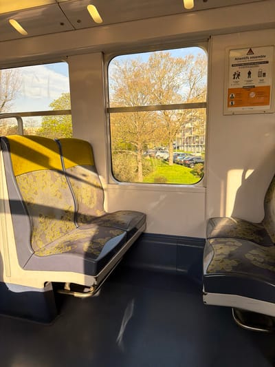 The image captures the interior of a public transport vehicle, likely a train or RER car, stopped or moving slowly in Arcueil, France. No individuals are present in the visible section of the carriage.
The interior features two empty passenger seats in the foreground, facing each other. The seat on the left is a double unit, upholstered in a grey-blue fabric with a repeating pattern of yellow, abstract, leaf-like shapes, and solid yellow headrests. The seat structure is light grey and dark blue. A single, matching seat is partially visible on the right. The floor is dark blue, while the upper walls and ceiling are white. Two oval-shaped interior lights are visible on the ceiling.
Between the two windows on the right side of the carriage, a white poster with a red triangular hazard symbol is displayed. The text "Attentifs ensemble" (Vigilant together) and "VEILLEZ A TOUTE SITUATION INHABITUELLE ?" (Pay attention to any unusual situation?) are clearly visible, accompanied by pictograms illustrating vigilance regarding luggage and suspicious behavior.
Through the windows, an exterior scene is visible under a clear blue sky with minimal cloud cover. Several deciduous trees with bare or nascent light-brown foliage stand prominently. Below the trees, a green, grassy slope descends to an area where multiple parked automobiles of various makes and colors are visible. In the background, behind the trees and parked vehicles, a multi-story building with numerous windows is discernible. Sunlight illuminates the exterior scene and casts distinct shadows within the train carriage.
